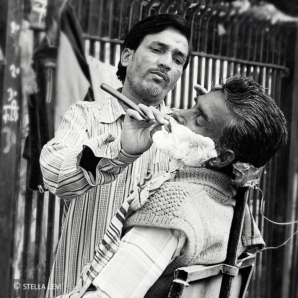Black-and-white photo of a street barber shaving a man in India